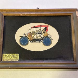 May include: Framed artwork featuring a vintage car design. The car is constructed from clock parts, with blue clock faces as wheels and a red canopy. The artwork is set against a black background within a wooden frame.