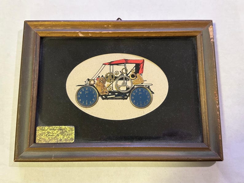 May include: Framed artwork featuring a vintage car design. The car is constructed from clock parts, with blue clock faces as wheels and a red canopy. The artwork is set against a black background within a wooden frame.