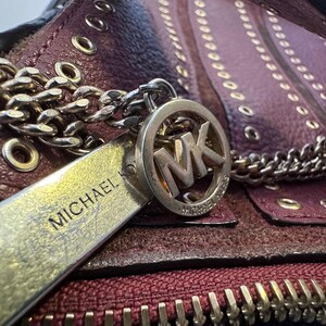 May include: Close-up of a burgundy leather handbag with a gold chain strap and zipper. The bag features a silver charm with the letters "MK" and the name "MICHAEL" engraved on a silver tag.
