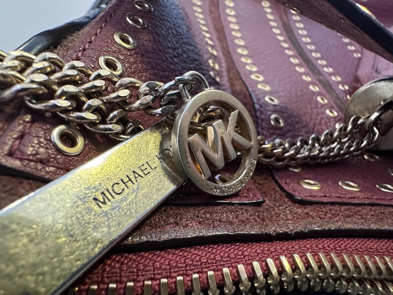 May include: Close-up of a burgundy leather handbag with a gold chain strap and zipper. The bag features a silver charm with the letters "MK" and the name "MICHAEL" engraved on a silver tag.