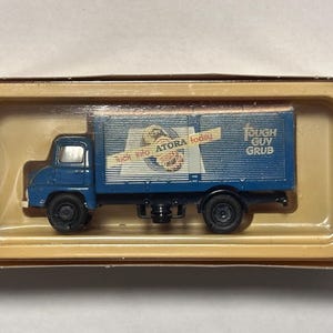 以下が含まれることがあります： 青いおもちゃのトラックで、箱型の荷台が付いています。トラックは黒い車輪と白いキャブが付いています。荷台には「Tough Guy Grub」と「Atora today」の文字があります。おもちゃは、茶色のプラスチック製のディスプレイケースに入っています。