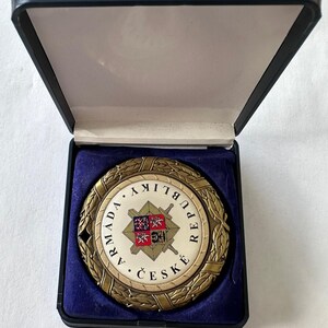 Op de afbeelding: Een ronde medaille in antieke stijl met de woorden "ARMADA REPUBLIKY ČESK&Eacute;" rond een centraal embleem. Het embleem toont een wapenschild met gekruiste zwaarden. De medaille wordt gepresenteerd in een zwart doosje met blauwe fluwelen voering.
