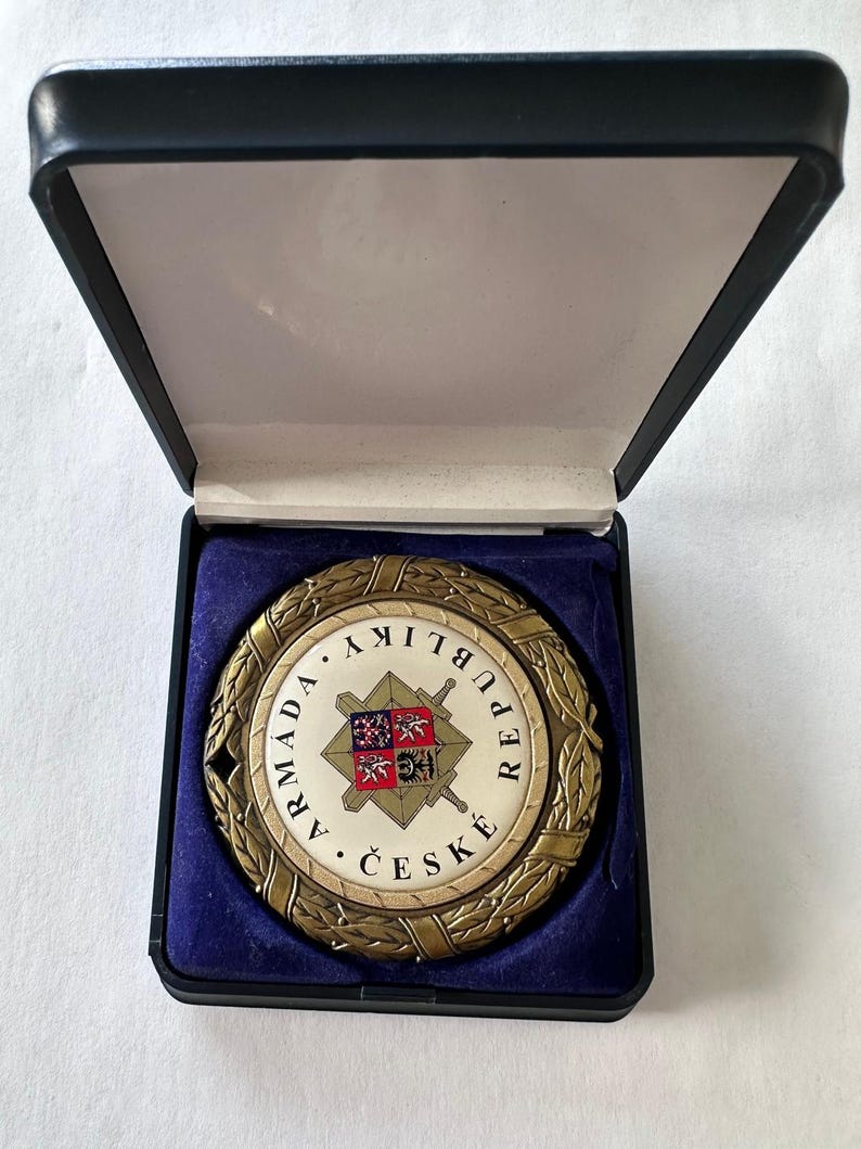 Op de afbeelding: Een ronde medaille in antieke stijl met de woorden "ARMADA REPUBLIKY ČESK&Eacute;" rond een centraal embleem. Het embleem toont een wapenschild met gekruiste zwaarden. De medaille wordt gepresenteerd in een zwart doosje met blauwe fluwelen voering.