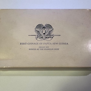 Pode incluir: Uma caixa retangular esbranqui&ccedil;ada com o texto "FIRST COINAGE OF PAPUA NEW GUINEA PROOF SET" e um emblema de p&aacute;ssaro e lan&ccedil;a. A caixa est&aacute; rotulada como "MINTED AT THE FRANKLIN MINT".