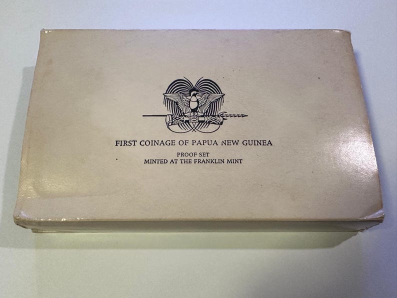 Pode incluir: Uma caixa retangular esbranqui&ccedil;ada com o texto "FIRST COINAGE OF PAPUA NEW GUINEA PROOF SET" e um emblema de p&aacute;ssaro e lan&ccedil;a. A caixa est&aacute; rotulada como "MINTED AT THE FRANKLIN MINT".