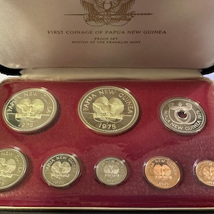 Pode incluir: Um conjunto de moedas da Papua Nova Guin&eacute; de 1975, apresentado numa caixa preta com interior de veludo vermelho. As moedas apresentam a inscri&ccedil;&atilde;o "Papua New Guinea" e o ano de 1975. A caixa est&aacute; rotulada como "First Coinage of Papua New Guinea".