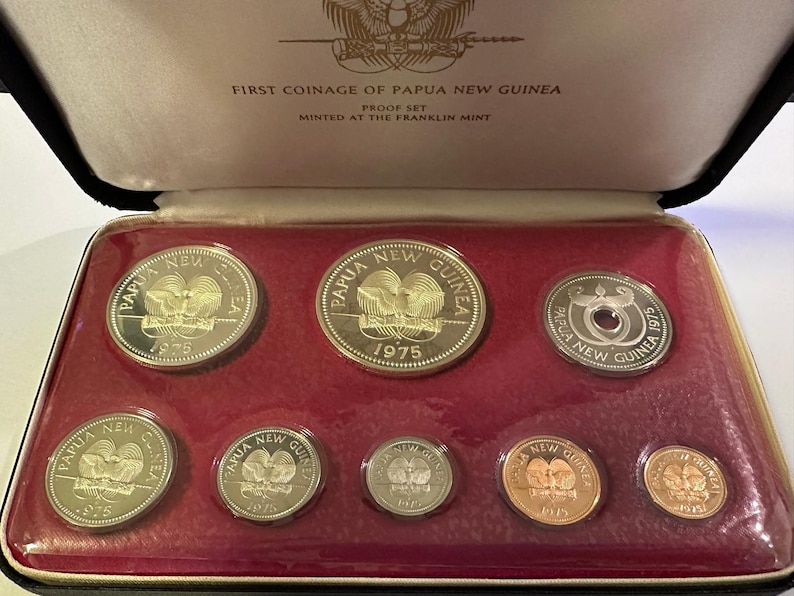 Pode incluir: Um conjunto de moedas da Papua Nova Guin&eacute; de 1975, apresentado numa caixa preta com interior de veludo vermelho. As moedas apresentam a inscri&ccedil;&atilde;o "Papua New Guinea" e o ano de 1975. A caixa est&aacute; rotulada como "First Coinage of Papua New Guinea".