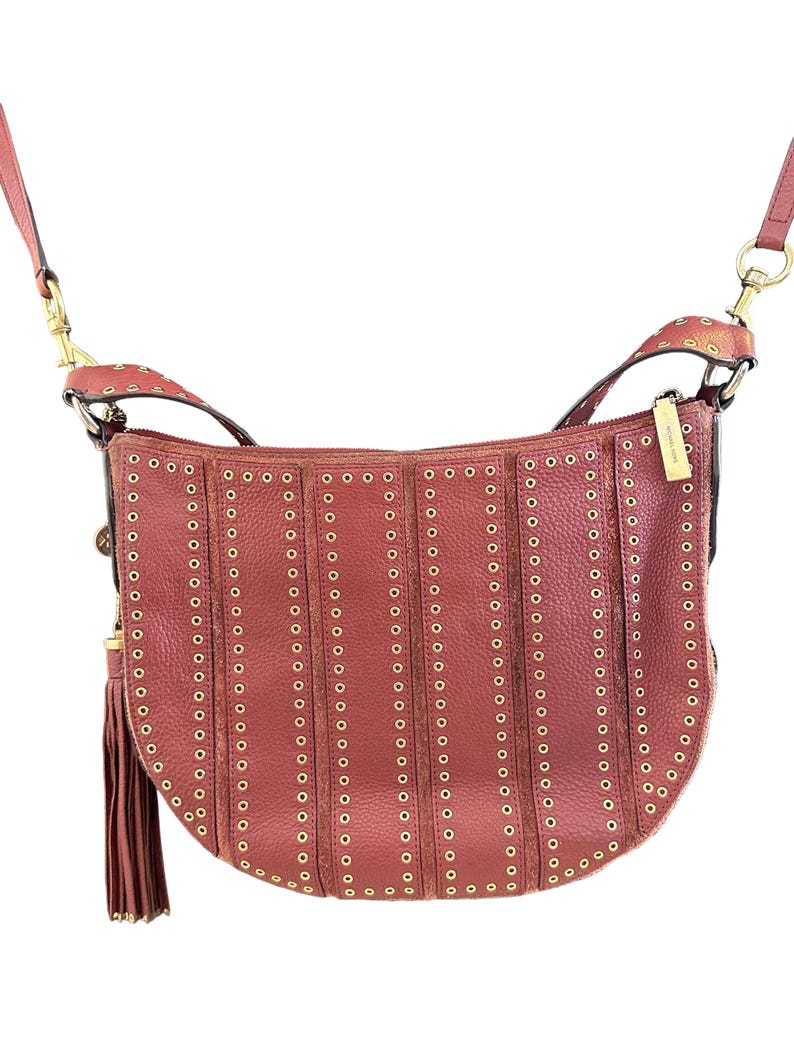 May include: A reddish-brown leather crossbody bag with gold-colored rivets and hardware. The bag has a curved shape, a long adjustable strap, and a tassel. The bag is a stylish accessory.