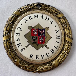 Op de afbeelding: Een cirkelvormige medaille in antieke stijl met een goudkleurige laurierkransrand. Het midden heeft een witte achtergrond met de woorden "ARM&Aacute;DA ČESK&Eacute; REPUBLIKY" in het zwart. Een centraal embleem bevat een schild met een wapen en gekruiste zwaarden.