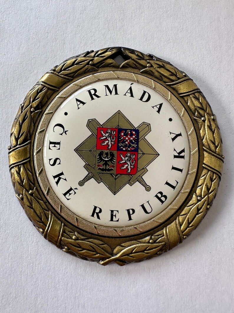 Op de afbeelding: Een cirkelvormige medaille in antieke stijl met een goudkleurige laurierkransrand. Het midden heeft een witte achtergrond met de woorden "ARM&Aacute;DA ČESK&Eacute; REPUBLIKY" in het zwart. Een centraal embleem bevat een schild met een wapen en gekruiste zwaarden.