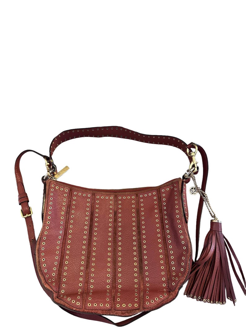 May include: A reddish-brown leather shoulder bag with gold-tone grommets and a matching tassel. The bag has a curved shape, a long adjustable strap, and a zipper closure. The bag is a stylish accessory.