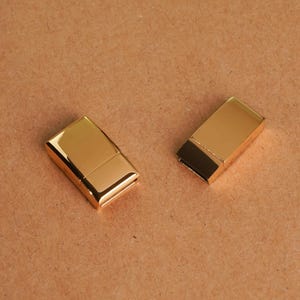 Pode incluir: Dois fechos magnéticos retangulares com acabamento dourado polido. Cada fecho consiste em duas peças interligadas com uma superfície lisa e reflexiva e bordas retas e limpas, projetadas para prender joias ou outros pequenos itens.