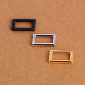 Könnte beinhalten: Drei rechteckige Metallschlaufen, die diagonal angeordnet sind. Die obere Schlaufe hat ein mattes schwarzes Finish, die mittlere Schlaufe ein poliertes silberfarbenes Finish und die untere Schlaufe ein poliertes goldfarbenes Finish. Jede Schlaufe hat abgerundete Ecken.