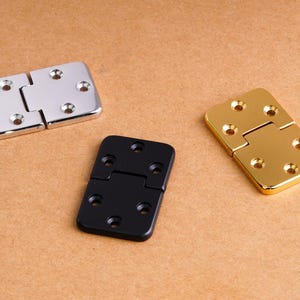 May include: Three rectangular metal hinges with rounded corners and six countersunk screw holes each. One hinge is polished silver, one is matte black, and one is polished gold. Each hinge features a central pin joint connecting two plates.
