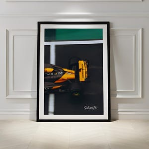 Peut inclure: Impression encadrée d'une voiture de course de Formule 1 en mouvement, principalement orange et noire. Le mot "Chrome" est visible. Le fond comprend une bande verte et la signature "Silverston".