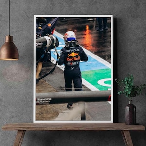 May include: A framed photograph of a Formula 1 race car driver in a Red Bull racing suit. The driver is walking on a wet track, with the Red Bull logo and sponsor details visible on the suit. The image is set against a backdrop of a pit lane.