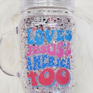 May include: Clear mason jar with a handle, filled with silver glitter and colorful confetti. The jar has the text "LOVES JESUS & AMERICA TOO" in blue and pink lettering.