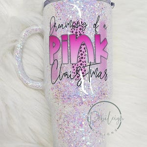 May include: Glitter-covered tumbler with a handle, featuring the text "Dreaming of a Pink Christmas" in pink and black. The tumbler has a pink leopard print design within the text and a silver lid.