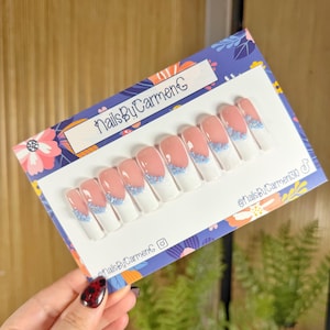 Pode incluir: Um conjunto de dez unhas postiças com um design de manicure francesa. As unhas apresentam uma ponta branca, uma base rosa e uma linha de pequenos enfeites azuis. Apresentadas num cartão com o texto "NailsByCarmenC".