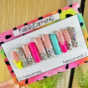 May include: A set of press-on nails with various designs. The nails feature a range of colors, including pink, yellow, and turquoise, with some adorned with rhinestones and leopard print accents. The text "NailsByCarmenG" is visible.