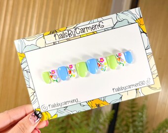 Puede incluir: Un conjunto de uñas postizas con un diseño floral. Las uñas alternan entre verde claro y azul, algunas con pequeños patrones de flores. La tarjeta dice "NailsByCarmen" e incluye identificadores de redes sociales.