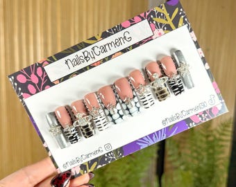 May include: A set of press-on nails featuring a variety of designs. Some nails are solid nude, while others have zebra, snake, and silver glitter patterns. Each nail is adorned with a silver charm. The card reads "NailsByCarmenG".