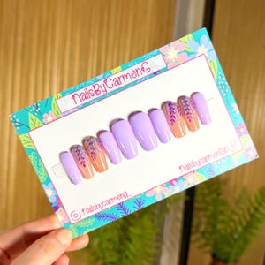 May include: A set of lavender-colored, coffin-shaped false nails. Some nails feature a design of orange and purple floral accents. The nails are displayed on a decorative card with the text "nailsbycarmeng" and floral patterns.