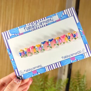 May include: A set of artificial nails with a floral design. The nails are painted with a nude base and feature colorful flowers in shades of pink, blue, yellow, and purple. The nails are displayed on a card with the text "NailsByCarmen".