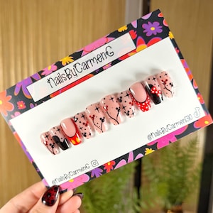 May include: A set of false nails with diverse designs. Some nails have a nude base with black floral and star patterns, while others have red and black accents. The set is displayed on a floral patterned card with the text "NailsByCarmenG".