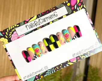 May include: A set of vibrant press-on nails featuring designs like smiley faces, lightning bolts, and chequered patterns. The nails are presented on a card with the text "NailsByCarmenG" and a floral design background.