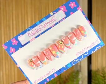 Puede incluir: Un conjunto de uñas postizas rosas con diseños de caras sonrientes, corazones, estrellas y arcoíris. Las uñas se muestran en una tarjeta azul con un patrón floral y el texto "NailsByCarmenC".