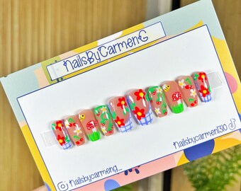 Puede incluir: Un conjunto de uñas postizas con base transparente y diseños florales coloridos. Cada uña presenta flores rojas, flores blancas, hojas verdes y un patrón de cuadrícula azul. El conjunto se muestra en una tarjeta con el texto "NailsByCarmenG".