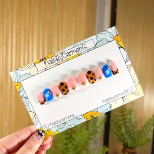 May include: A set of press-on nails with a variety of designs. The nails feature a combination of pink, white, blue, and leopard print patterns. Some nails have gold star and gem accents. The card reads "NailsByCarmenC".