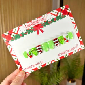 Op de afbeelding: Een set kunstnagels met een kerstthema. De nagels hebben ontwerpen van de Grinch, rood-witte strepen en glitter. De nagels worden gepresenteerd op een kaart met een rood-groene geruite rand en de tekst "NailsByCarmenG".