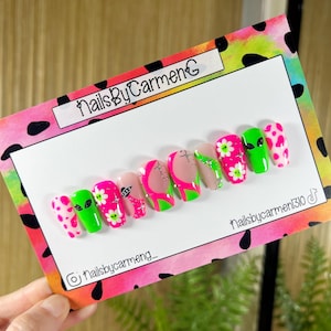 May include: A set of press-on nails with a vibrant, colorful design. The nails feature alien and flower graphics, along with pink and green accents. The card reads "NailsByCarmenG" and includes social media handles.