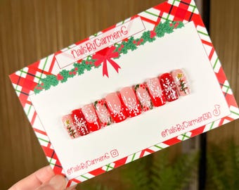 May include: A set of Christmas-themed press-on nails. The nails feature red, white, and pink designs, including snowflakes and Christmas trees. The nails are displayed on a card with a red and green plaid pattern and the text "NailsByCarmenG".