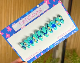 Puede incluir: Un conjunto de uñas postizas turquesas con diseños azul oscuro y adornadas con pedrería y pequeñas flores azules. Las uñas se exhiben en una tarjeta azul y blanca con el texto "NailsByCarmenC".