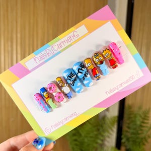 Puede incluir: Un conjunto de uñas postizas de diseño personalizado con personajes de Los Simpson. Las uñas están pintadas con colores vibrantes e incluyen imágenes de Homer, Bart, Lisa y otros personajes, junto con el logotipo del programa y rosquillas.