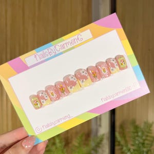 May include: A set of press-on nails with a floral design. The nails feature a light pink base with white flowers, yellow accents, and framed floral illustrations. The card reads "NailsByCarmenC" and includes social media handles.
