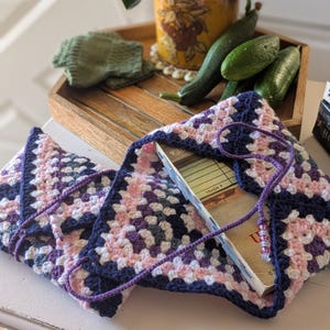 May include: A crocheted book bag in a granny square pattern, featuring pink, white, purple, and navy blue squares. The bag has a matching navy blue border and a purple drawstring with decorative beads. A book is inside the bag.