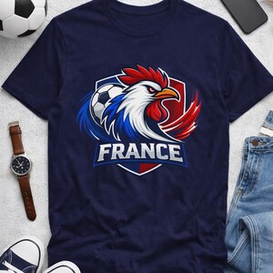 May include: Navy blue t-shirt featuring a rooster design with the word "FRANCE" in white. The rooster is in the colors of the French flag, with a soccer ball. A watch, phone, shoes, and jeans are also in the image.