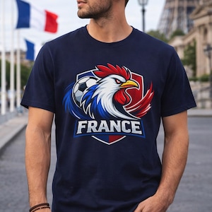May include: Navy blue t-shirt featuring a graphic of a rooster, soccer ball, and the word "FRANCE" in white. The rooster has red, white, and blue feathers. The Eiffel Tower and French flags are in the background.