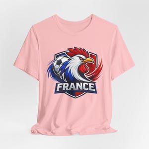 Może przedstawiać: Jasnor&oacute;żowa koszulka z grafiką koguta i piłki nożnej w barwach francuskiej flagi. Słowo "FRANCE" jest wyświetlane w niebieskim banerze pod kogutem.