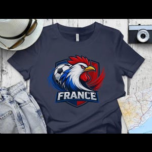 Może przedstawiać: Granatowa koszulka z grafiką koguta i piłki nożnej, z napisem "FRANCE". Kogut jest czerwono-biało-niebieski, z ż&oacute;łtym dziobem. Projekt znajduje się na tle w kształcie tarczy.