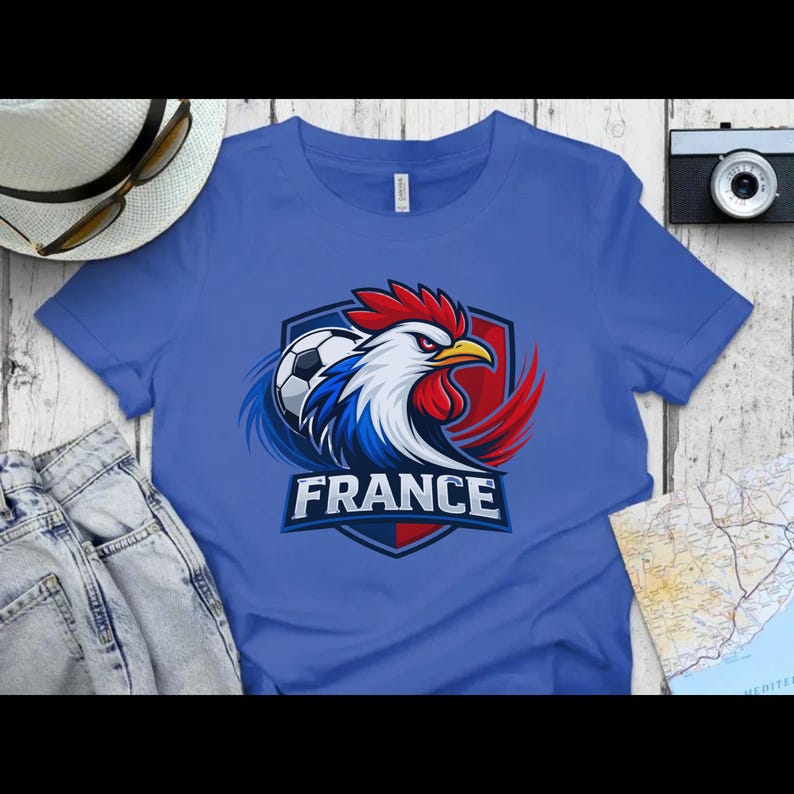 Może przedstawiać: Niebieska koszulka z grafiką koguta, piłki nożnej i napisem "FRANCE". Kogut jest w kolorach czerwonym, białym i niebieskim, czyli barwach francuskiej flagi. Projekt znajduje się na niebieskiej koszulce.