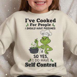 Könnte beinhalten: Ein cremefarbenes Sweatshirt mit dem Text "I've Cooked For People I SHOULD HAVE POISONED." Darunter befindet sich eine Cartoon-Figur des Grinch mit Kochmütze, die in einem Topf rührt. Der Text "SO YES, I DO HAVE Self Control" steht darunter.