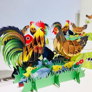 May include: A 3D paper art scene featuring a rooster and hen with chicks, set on a green grassy base. The rooster has a large, colorful tail. Other chickens and farm elements are in the background. The artwork is a pop-up style.