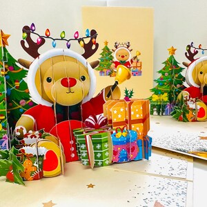 Könnte beinhalten: Eine festliche Pop-up-Weihnachtskarte mit einem Rentier in einem Weihnachtsmann-Anzug und einer Lichterkette. Die Karte enthält einen Weihnachtsbaum und Geschenke. Der Hintergrund enthält eine zweite Karte mit einem Rentier und einem Weihnachtsbaum.