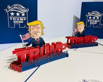 May include: Red and blue 3D pop-up cards with the word "TRUMP" in large letters. The cards feature a cartoon of a man holding a small American flag. The phrase "MAKE AMERICA GREAT AGAIN" is also visible.