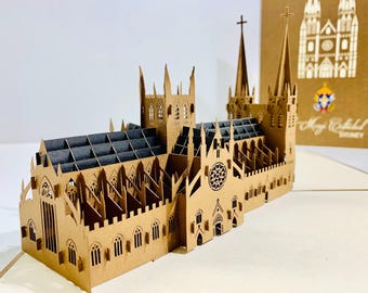 Puede incluir: Una maqueta de papel tridimensional detallada de una catedral, con intrincadas ventanas recortadas y un techo gris oscuro. El modelo está hecho de papel de color beige e incluye dos altas agujas con cruces. Una tarjeta de felicitación con el texto "Mary Cathedral SYDNEY" está en el fondo.